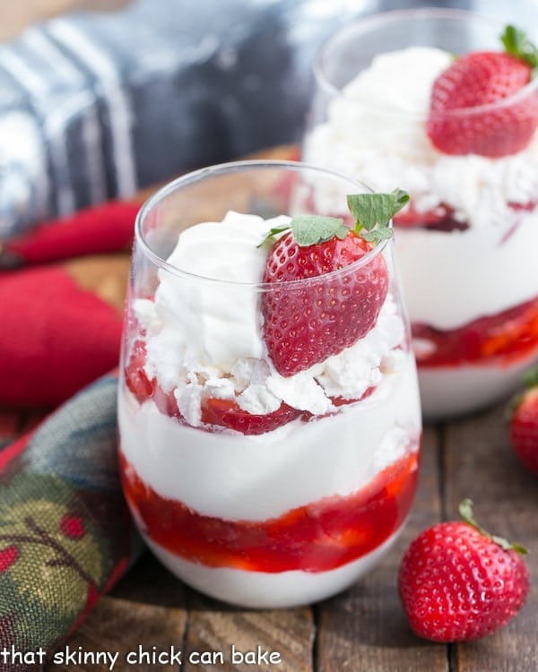 Fruit parfait with layers of whipped cream, crushed meringue cookies and sliced strawberries in parfait glasses with fresh strawberries nearby