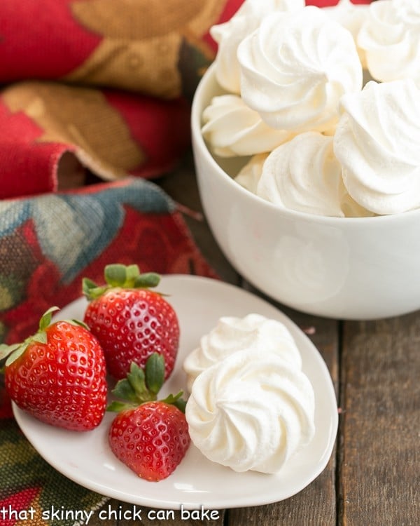 Egg White cookies are incredibly light, ethereal cookies made from whipped egg whites and sugar and will melt in your mouth and tantalize your sweet tooth! This easy meringue cookie recipe will become one of your favorites. #cookies #recipe #holidaybaking white bowl filled with Simple Meringue Cookies a plate of cookies and fresh strawberries sits next to it.