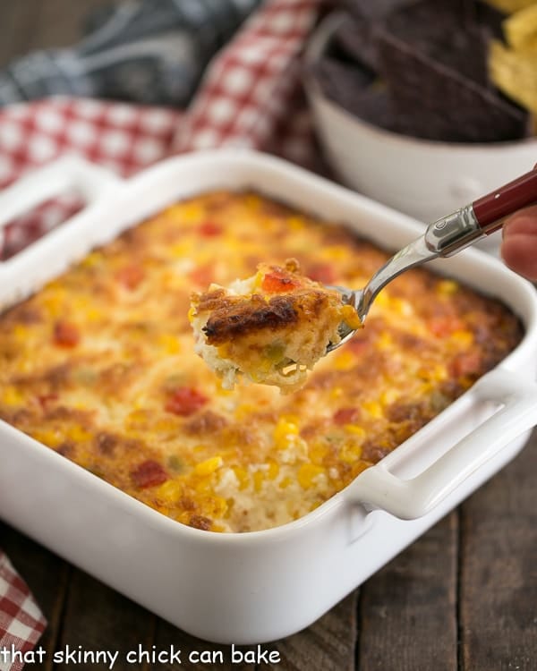Fiesta Corn Dip - With a double dose of gooey cheese, you'll want to serve this magnificent dip at every gathering! A spoonful of Fiesta Corn Dip on a red handled serving spoon with a white casserole in the background.