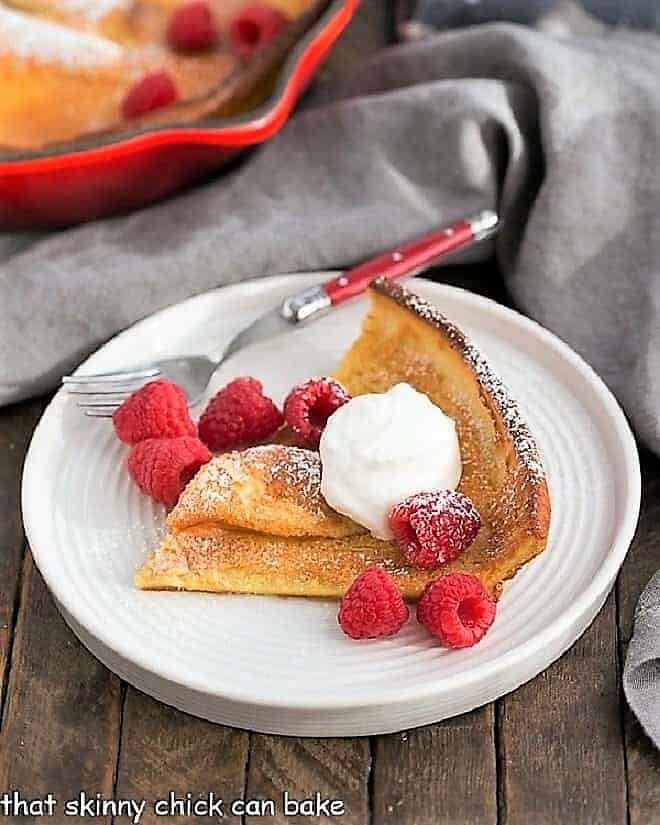 Best Dutch Baby or German Pancake Recipe - A puffed breakfast dish topped with berries, whipped cream and powdered sugar! Best Dutch Baby Pancake slice on a white plate topped with cream and raspberries.