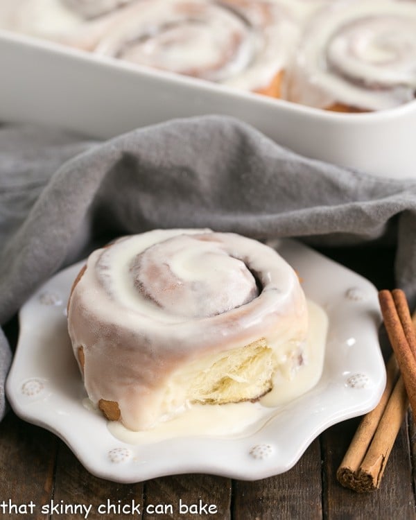 A cinnamon bun on a plate with the pan of rolls in the background