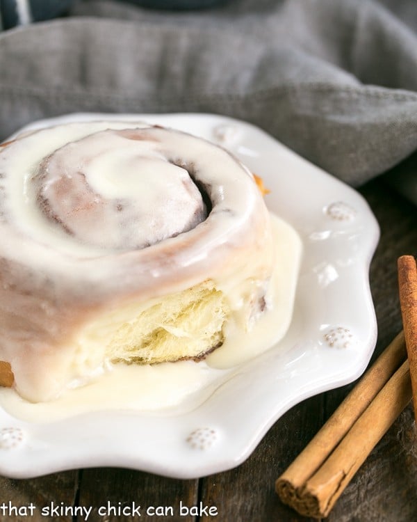 A glazed cinnamon bun on a white plate with cinnamon stick garnishes