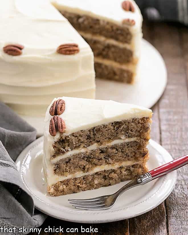 Hummingbird Cake with Coconut slice on a white dessert plate with the whole cake in the background.