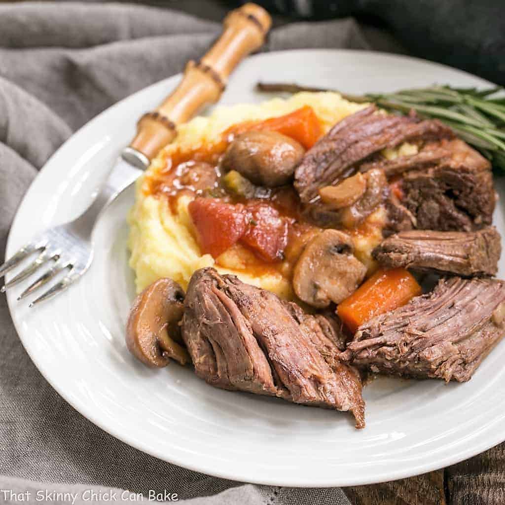 Pot Roast in Oven with Mushrooms, Tomatoes & Red Wine on a white ceramic plate with a bamboo handled fork.