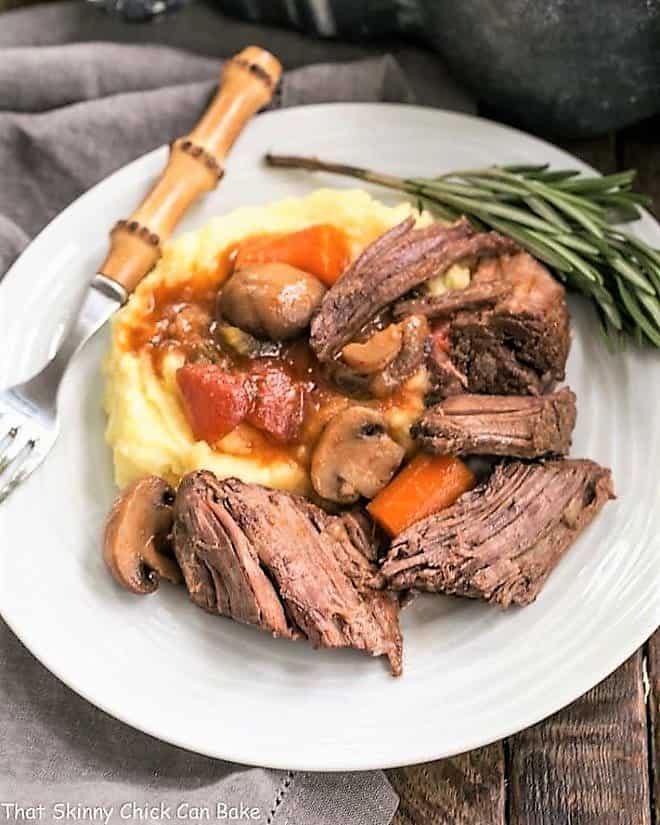 Pot Roast with Mushrooms, Tomatoes & Red Wine on a white plate with mashed potatoes and a sprig of rosemary.