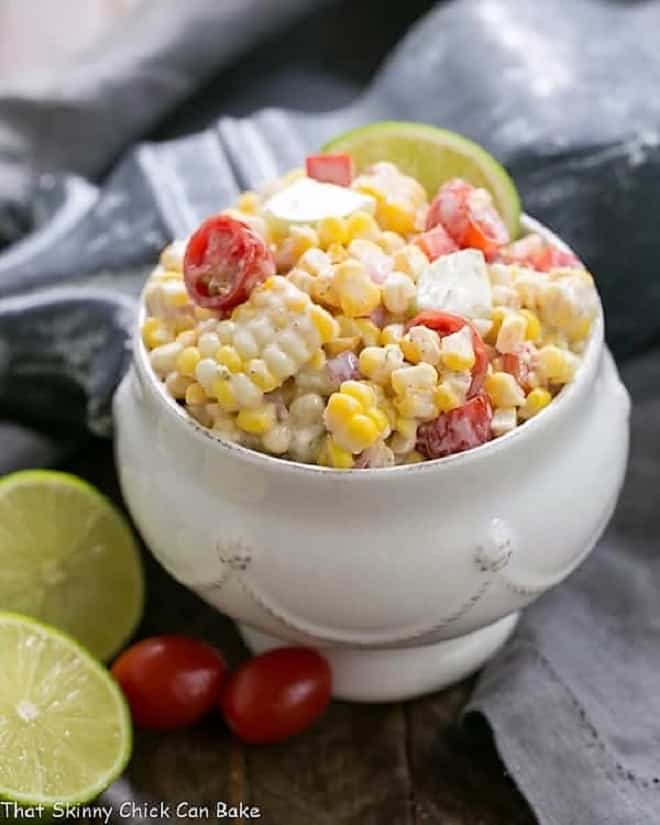 Spicy Mexican Street Corn Salad garnished with a lime slice in a white bowl Spicy Mexican Street Corn Salad in a white tureen with limes and tomatoes.