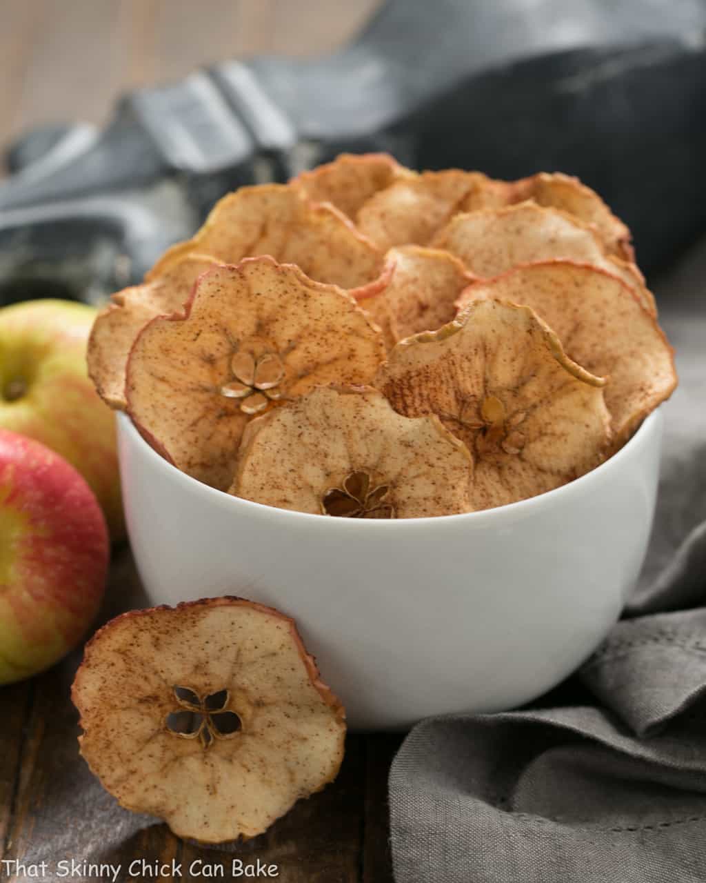 Cinnamon Apple Chips in a white ceramic bowl next to fresh apples.