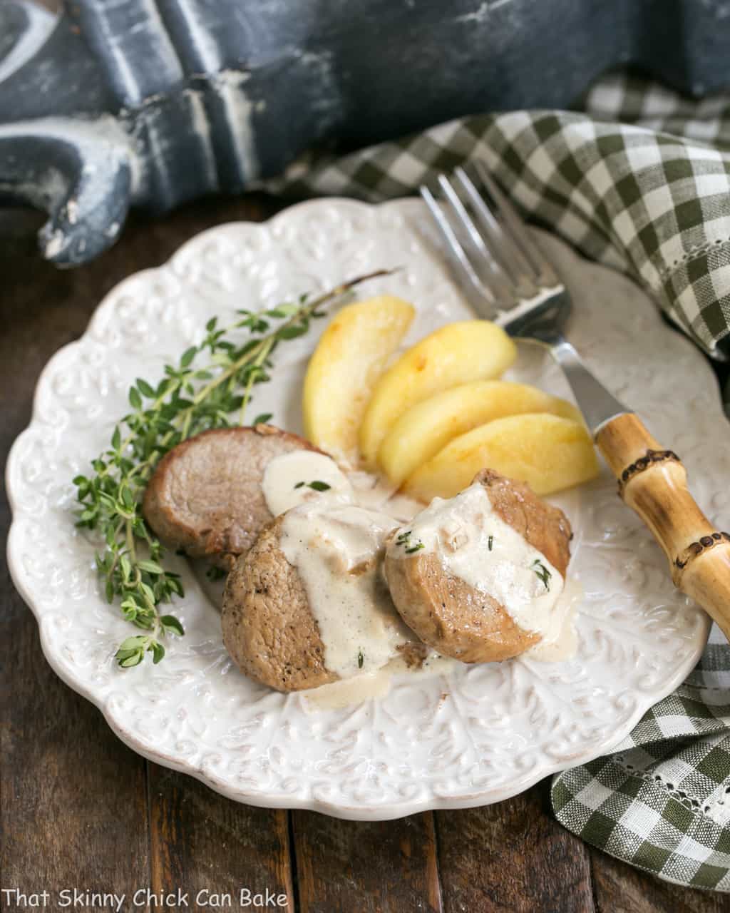 Apple Topped Pork Medallions with Calvados Cream Sauce on a dinner plate with fresh thyme and a bamboo handle fork.