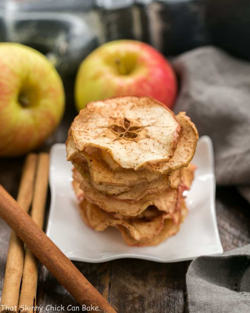 A stack of Cinnamon Apple Chips on a square white plate with fresh apples and cinnamon sticks