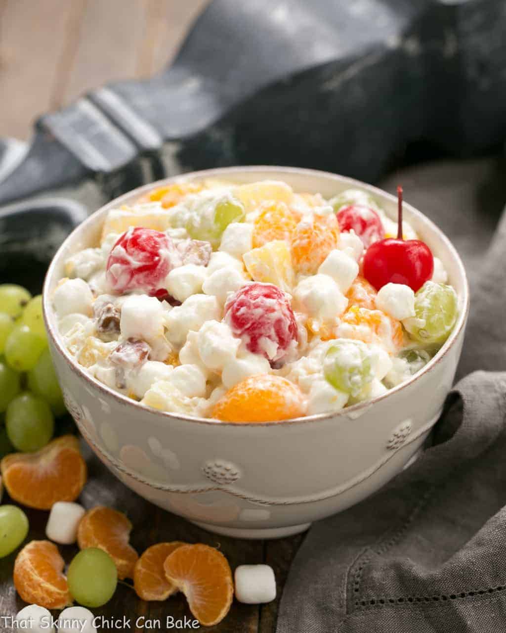 The Best Ambrosia Salad in a white serving bowl surrounded by fruit and marshmallows.