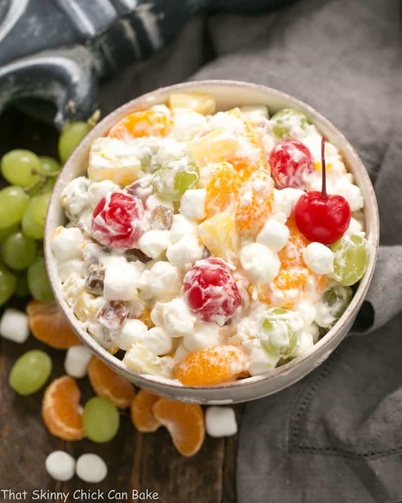 The Best Ambrosia Salad {No Cool Whip} in a white ceramic bowl surrounded by grapes, marshmallows and orange segments