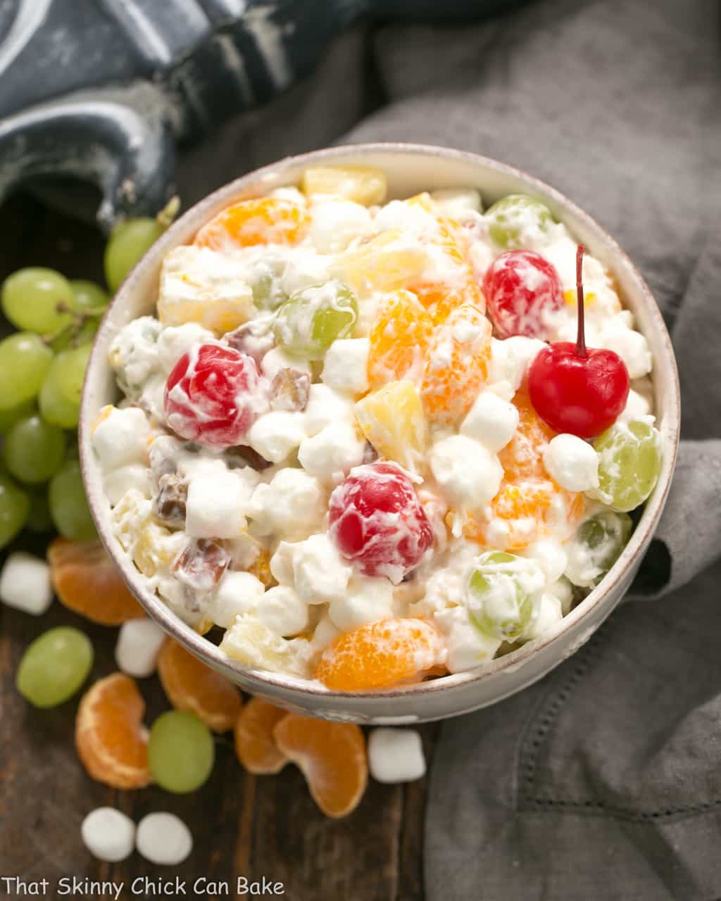 Ambrosia Salad in a white ceramic bowl surrounded by grapes, marshmallows and orange segments.