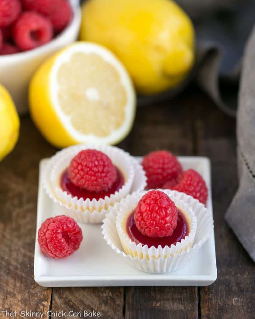 Two raspberry topped cheesecake bites on a small white plate with raspberry garnishes