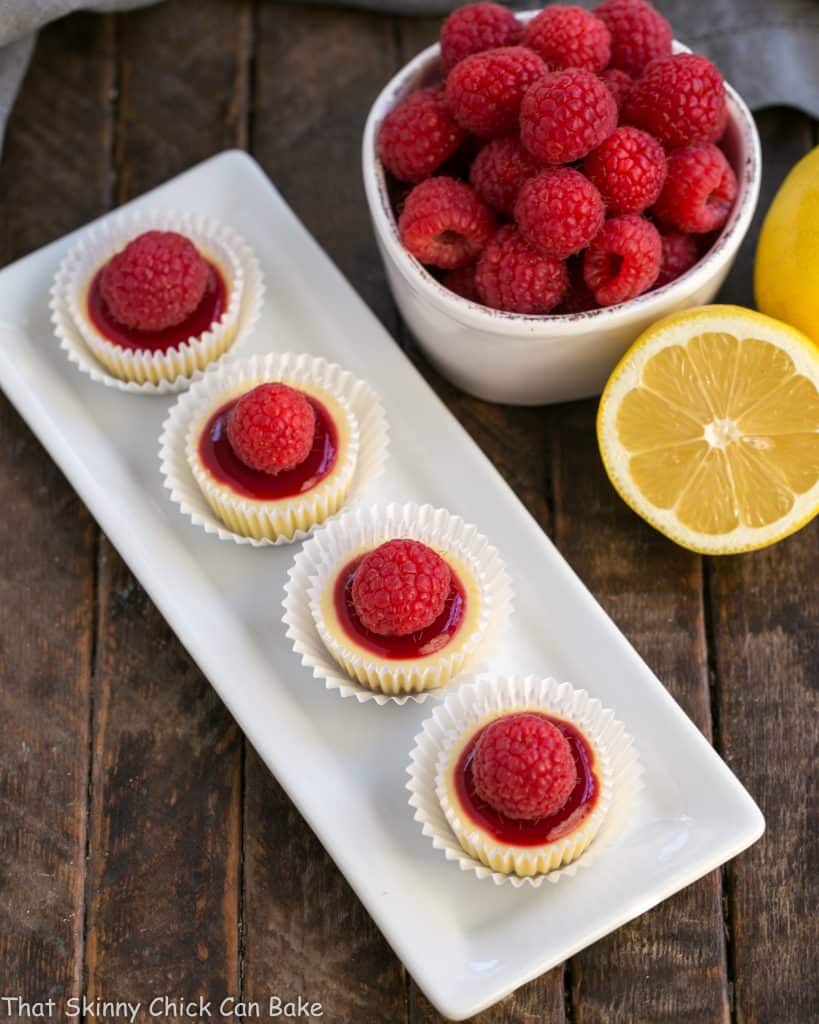 Overhead view of a white dessert platter with bite sized cheesecakes topped with raspberry sauce and fresh raspberries