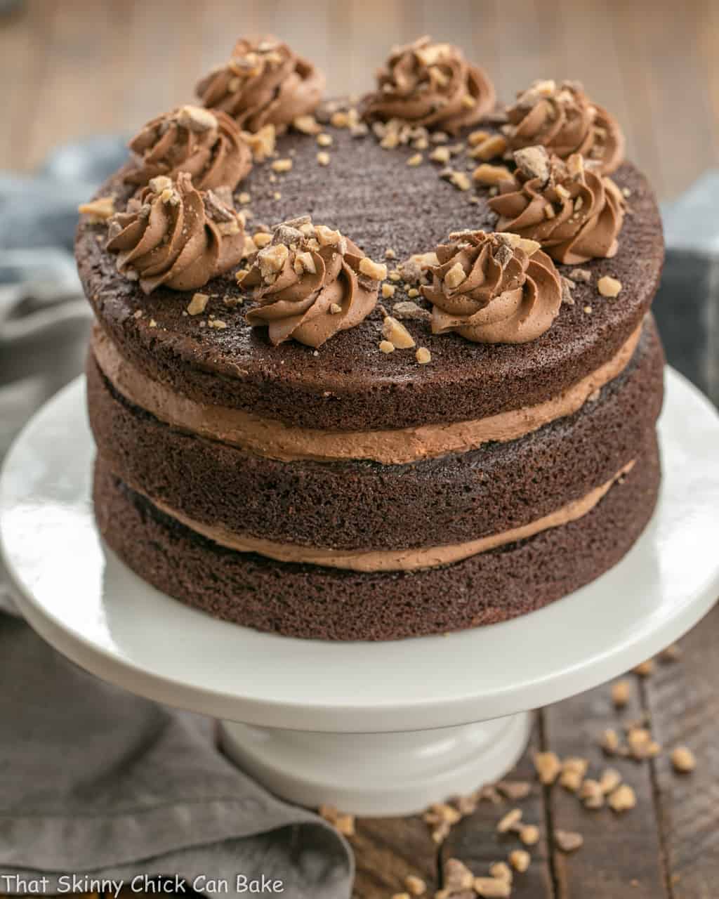 Triple Layer Chocolate Toffee Cake - 3 rich chocolate layers filled with chocolate buttercream and toffee bits Full side view of a Triple Layer Chocolate Toffee Cake on a white ceramic cake stand.