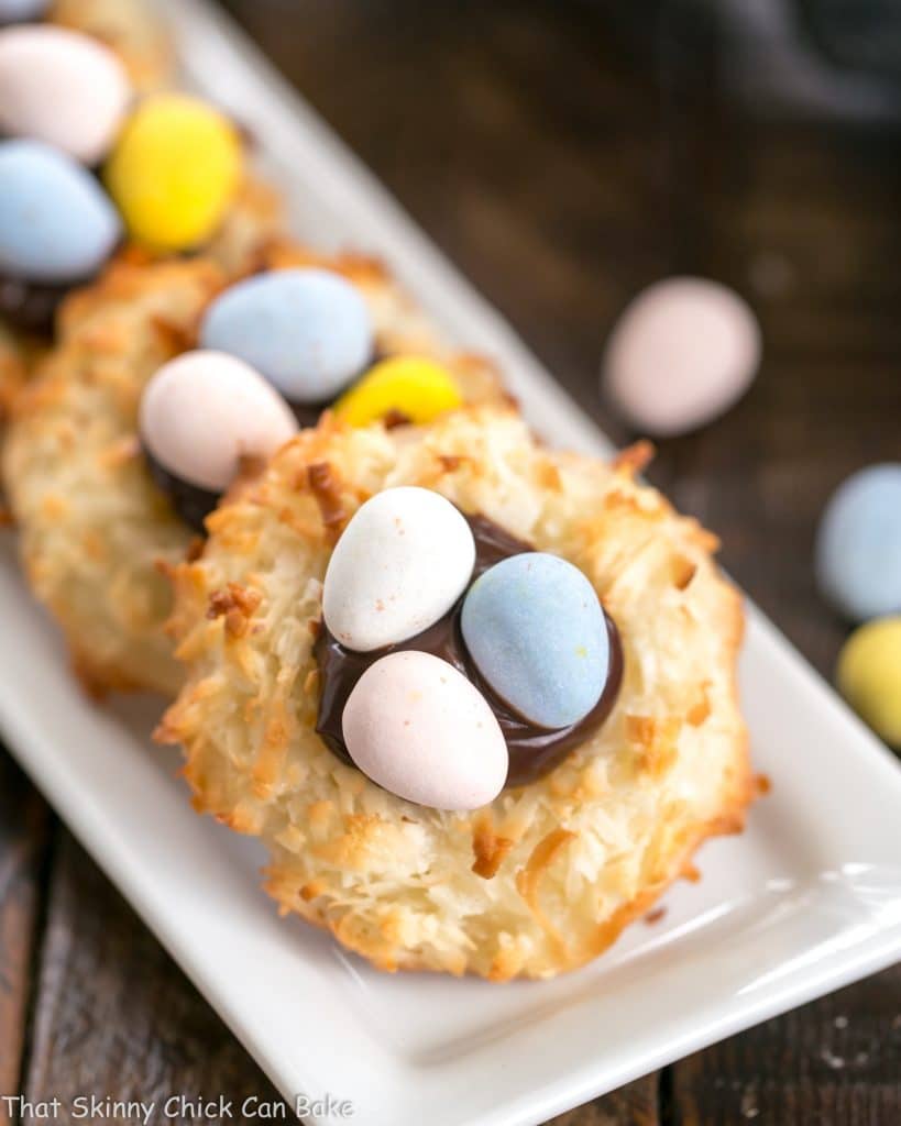 Coconut Macaroon Easter Nests filled with fudge sauce and topped with chocolate eggs! A delicious Easter cookie recipe! Coconut Macaroon Easter Nests lined up on a thin white tray