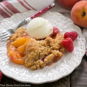 Peach raspberry crisp on a decorative white dessert plate topped with a scoop of ice cream and garnished with 2 raspberries