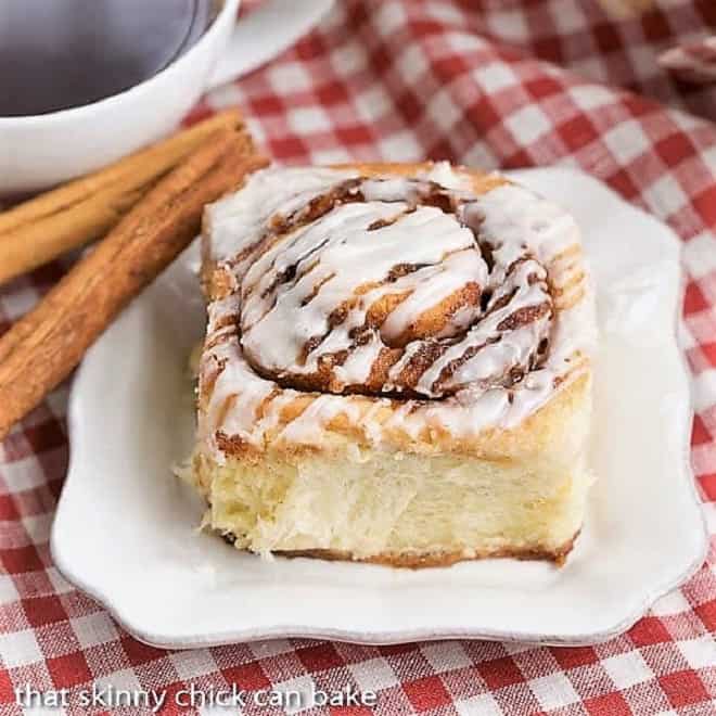 The Best Cinnamon Buns on a square white plate with cinnamon sticks on a red and white checked napkin.