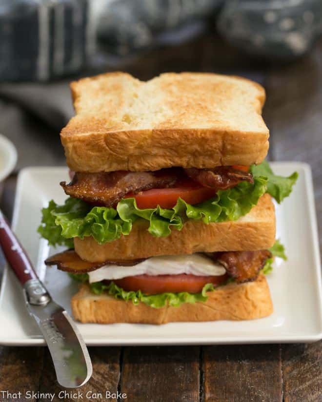 A double decker Brie Bacon Lettuce and Tomato Sandwich on a square white plate with a small red handled knife.