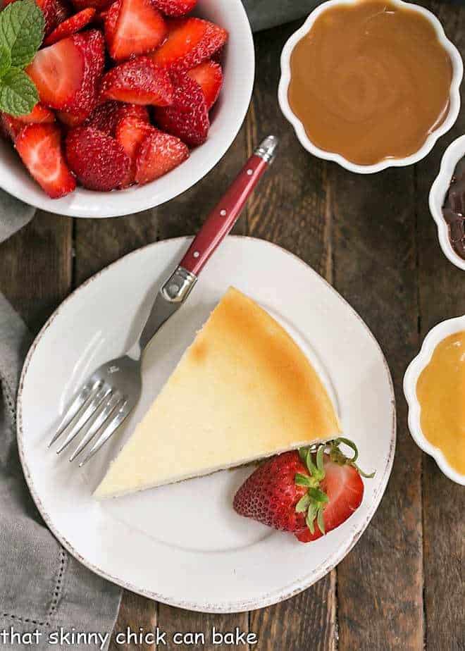 Overhead of perfect vanilla cheesecake with a bowl of strawberries and toppings.