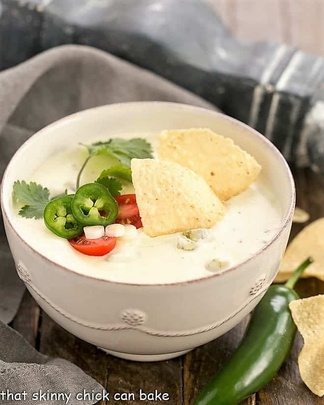 Easy Homemade Queso Dip in a white serving bowl topped with tortilla chips, jalapeno slices, cilantro sprig.