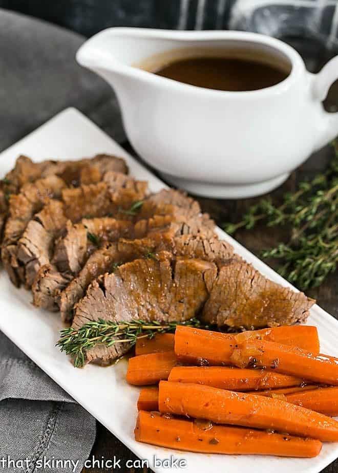 Braised Cola Brisket with Bourbon Gravy on a white platter with carrots and a pitcher of gravy.