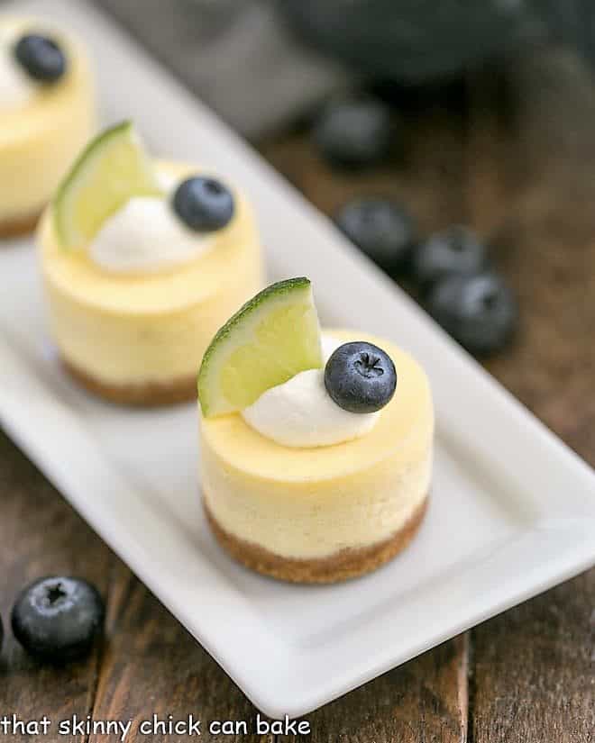 Mini Margarita Cheesecakes topped with cream, a lime slice and a blueberry on a white tray