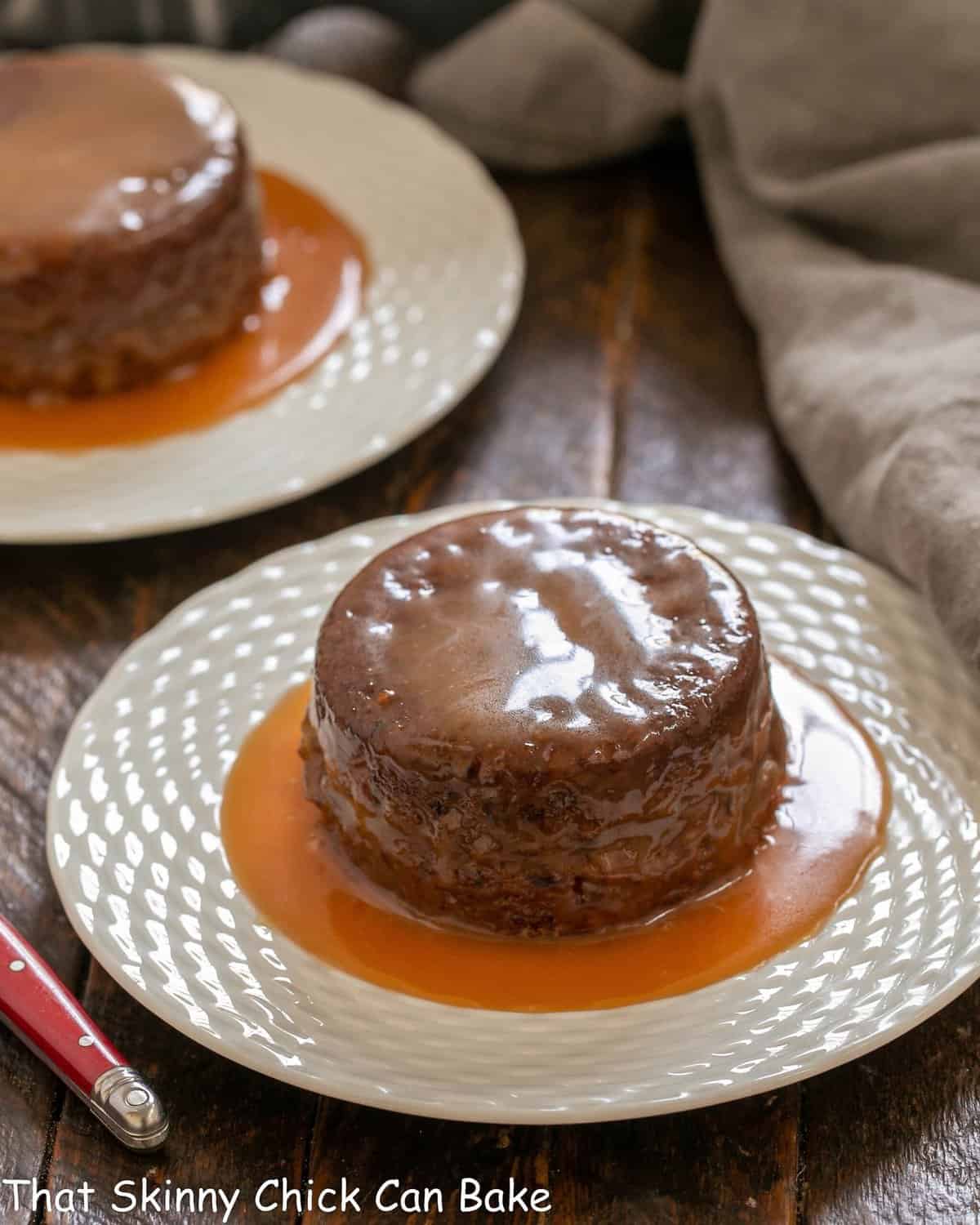 Sticky toffee pudding cakes on two white dessert plates surrounded by caramel sauce.