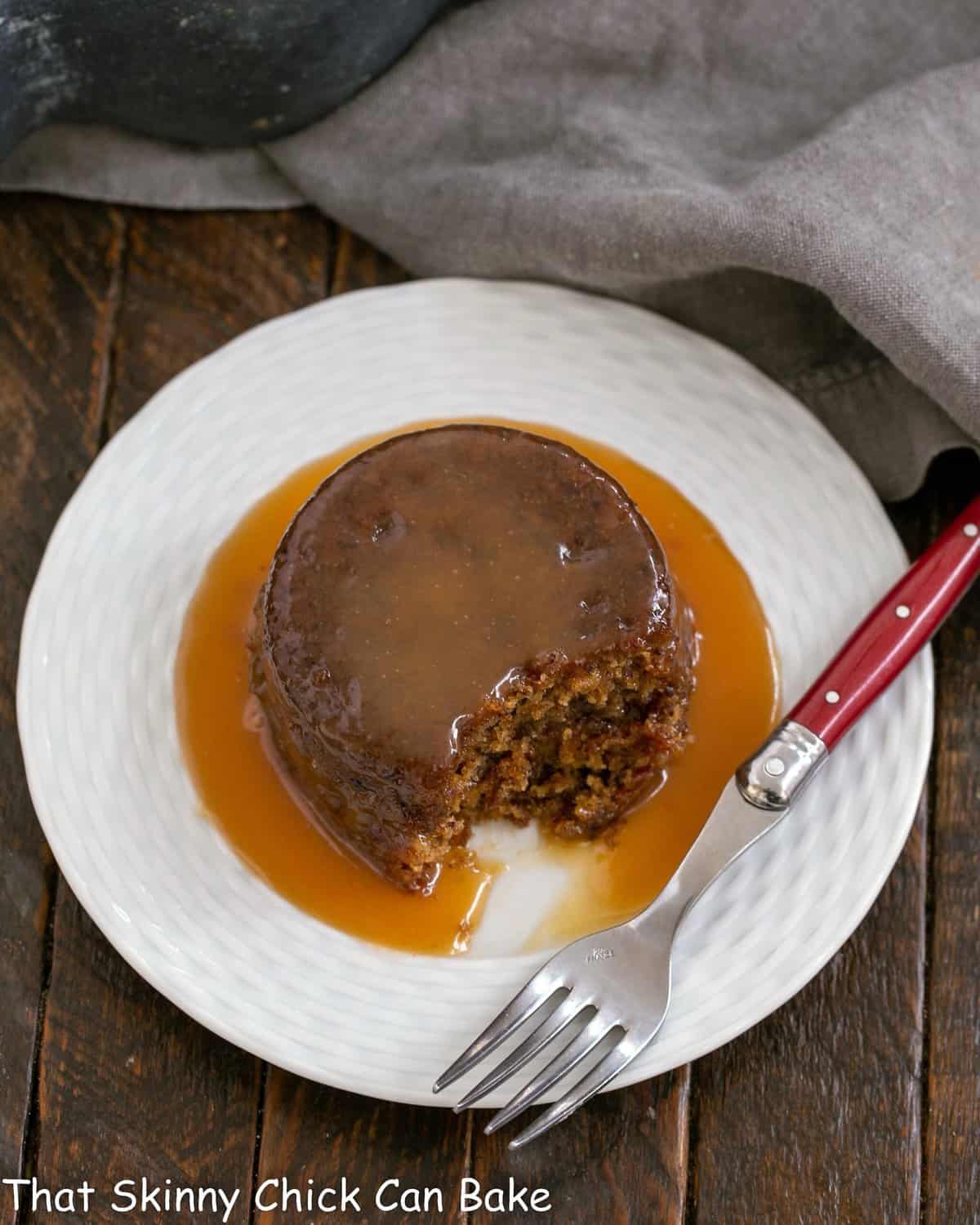 Overhead view of sticky toffee pudding on a white dessert plate with a red handle fork.