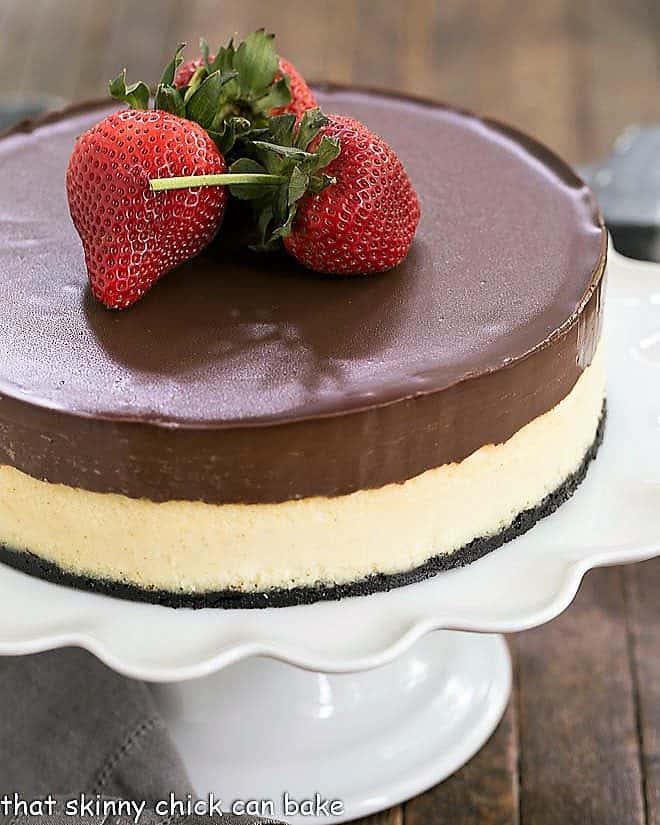 Ganache Topped Cheesecake on a white cake stand topped with large strawberries