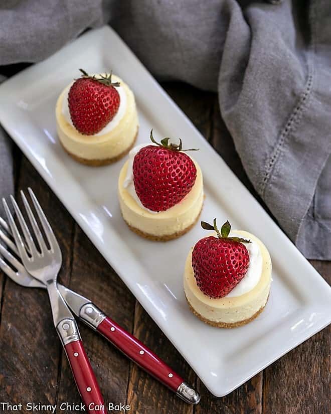 Overhead view of mini vanilla bean cheesecakes on a white platter with 3 red handled forks