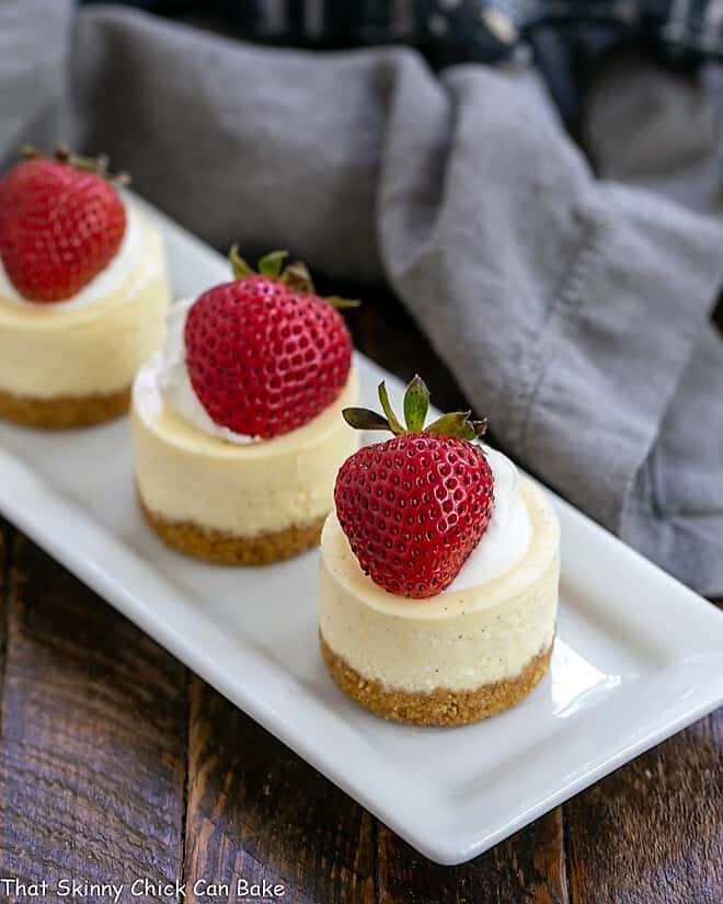 Vanilla Bean Mini Cheesecakes topped with strawberries and cream on a white platter.