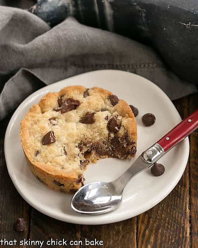 Round chocolate chip cookie bar with a few bites removed on a round white plate with a red handled spoon