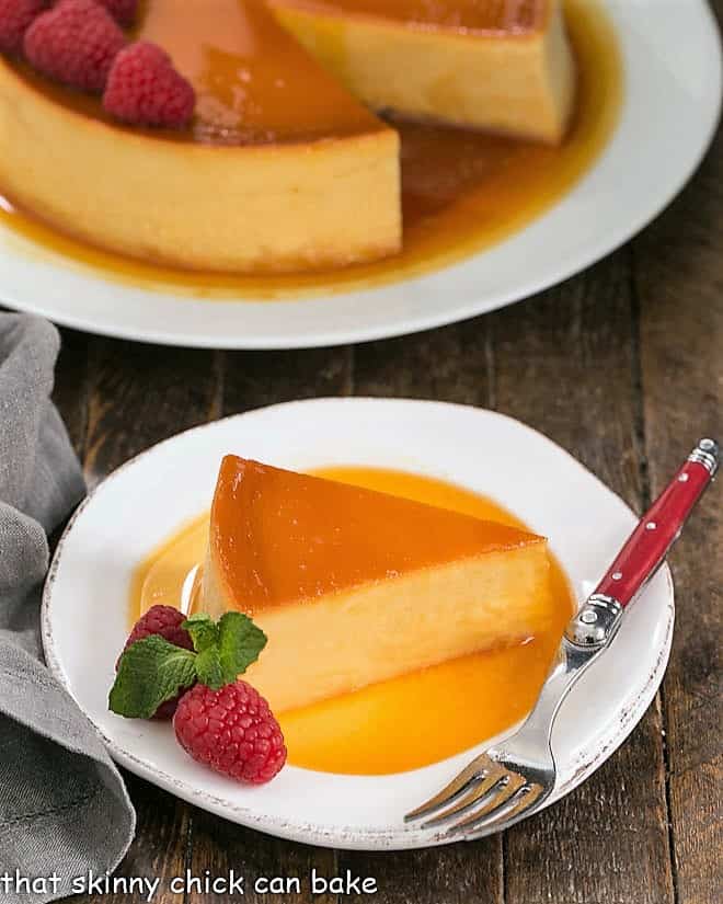 This rich and dreamy Caramel Flan Recipe is a cross between creme brulee and cheesecake! Everyone will want to lick their plates clean! Slice of Leche Flan on a white plate with raspberry garnish.