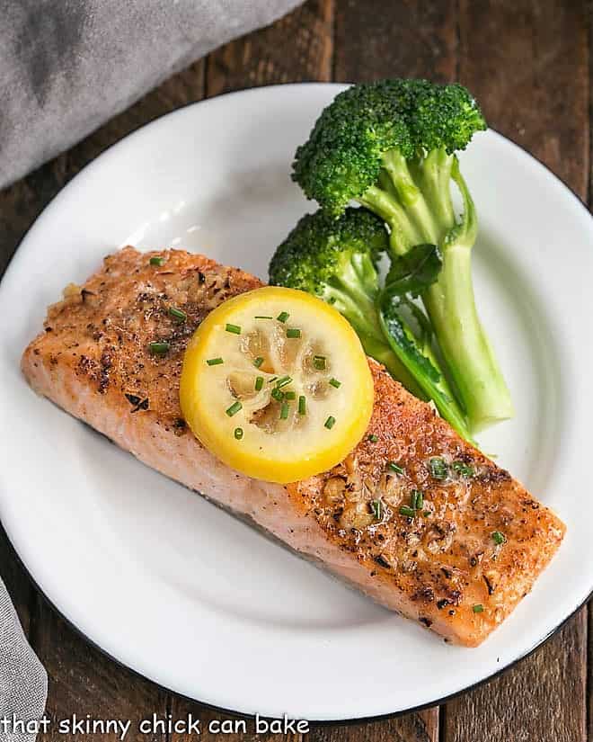 Overhead view of Salmon fillet topped with a lemon slice on a white plate with broccoli.