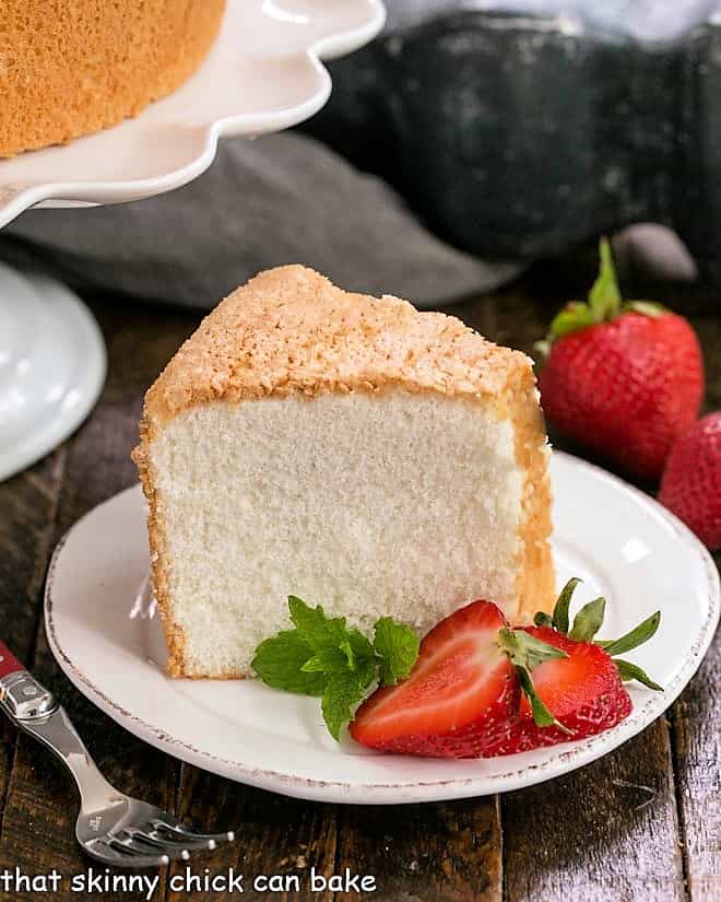 Slice of angel food cake on a white dessert plate with sliced strawberries and mint.