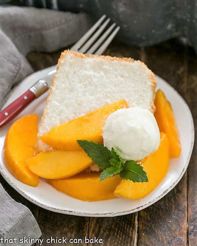 A slice of angel food cake on a dessert plate topped with fresh peach slices, whipped cream and mint.