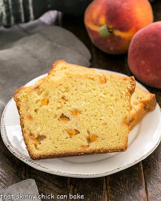 Two slices of peach cake on a white dessert plate with 2 peaches in the background.