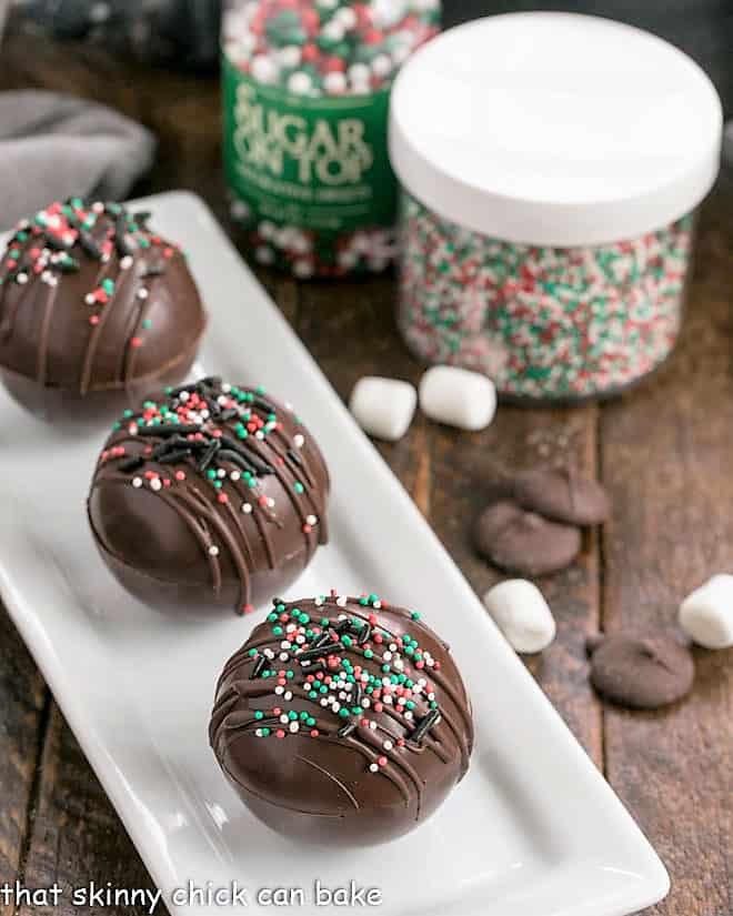 3 hot chocolate bombs on a white tray with sprinkles, marsmallows and hocolate melts.