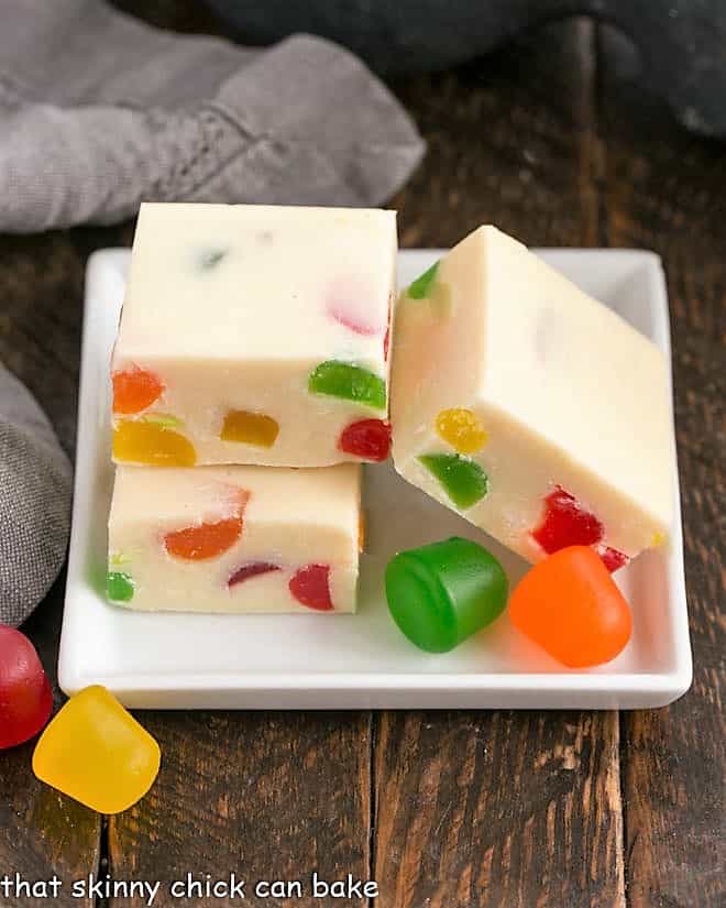 3 cubes of festive white chocolate fudge on a square white plate garnished with gumdrops.