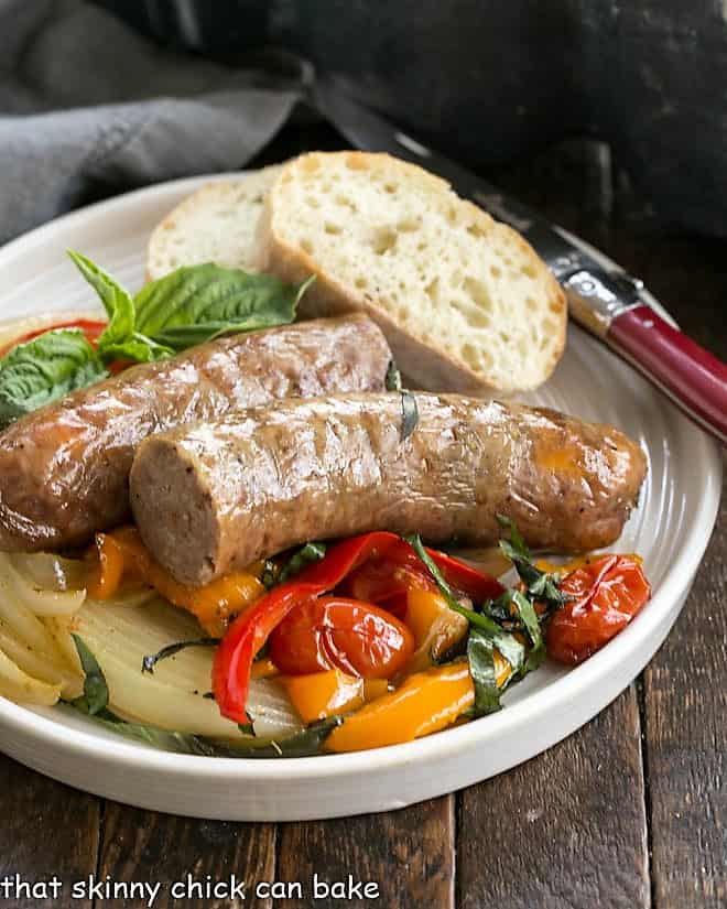 Sheet Pan Sausage and peppers served on a white dinner plate with a slice of rustic bread and a red handled fork