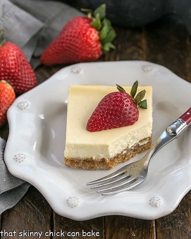 One cheesecake bar topped with a half strawberry on a decorative white plate with a fork