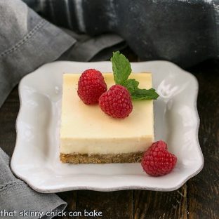 One vanilla cheesecake bar on a white plate topped with raspberries and a sprig of mint
