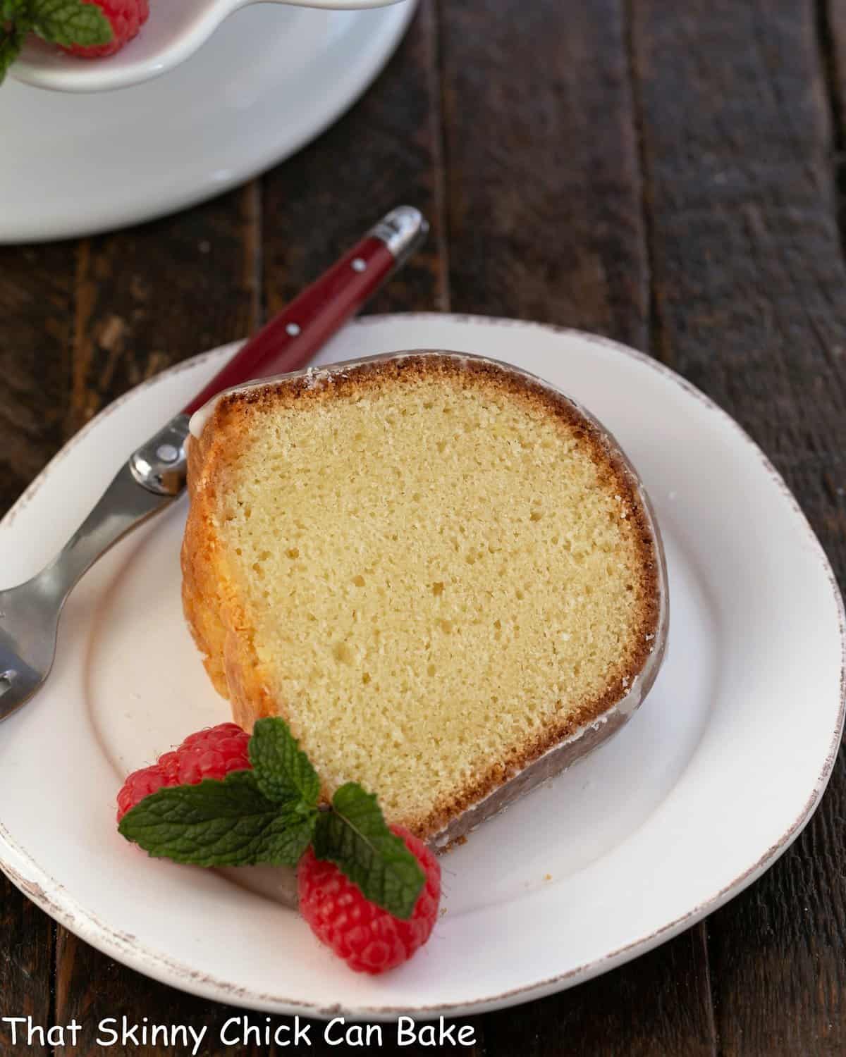 A slice of sour cream pound cake on a white desssert plate with a red handle fork and fresh raspberries.