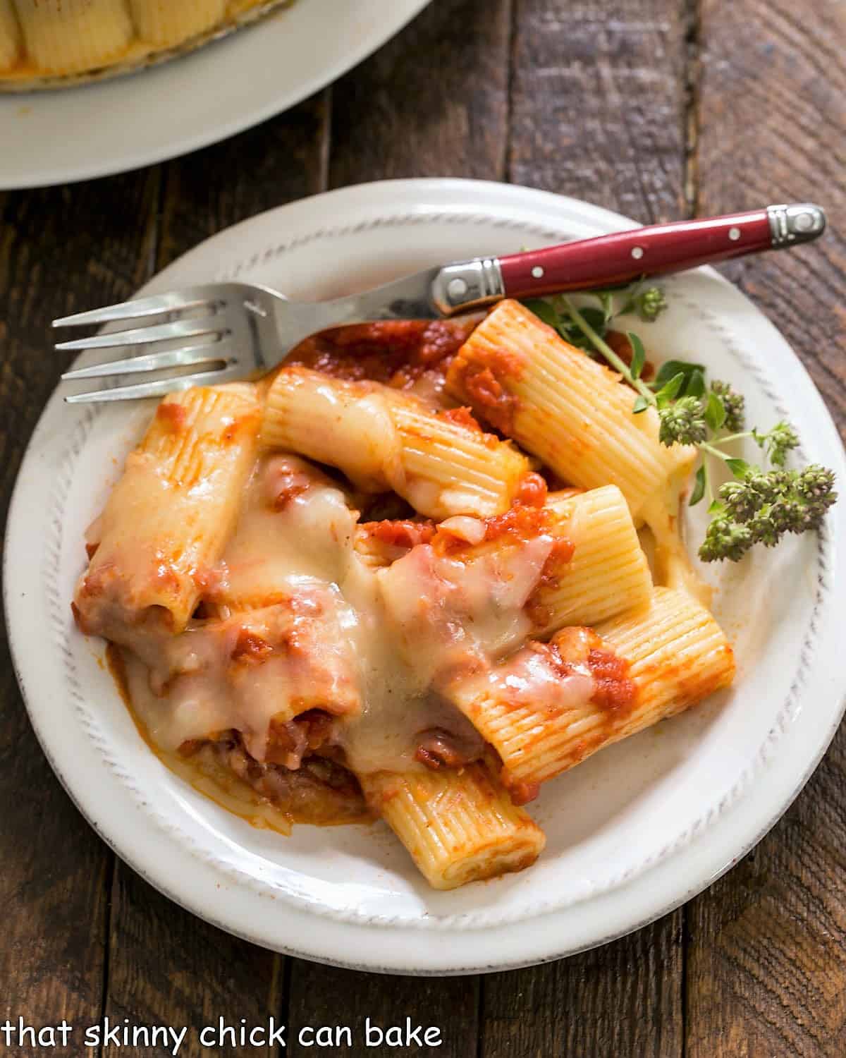 Overhead view of baked rigatoni on a small wite plate with a fork and herb garnish.