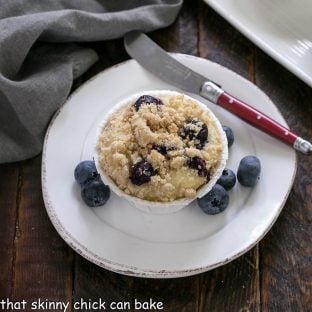 Overhead view of streusel topped blueberry muffin on a round white plate garnished with fresh blueberries and flanked with a red handle knife