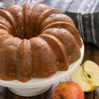 Apple Bundt Cake on a white cake stand next to 2 apples