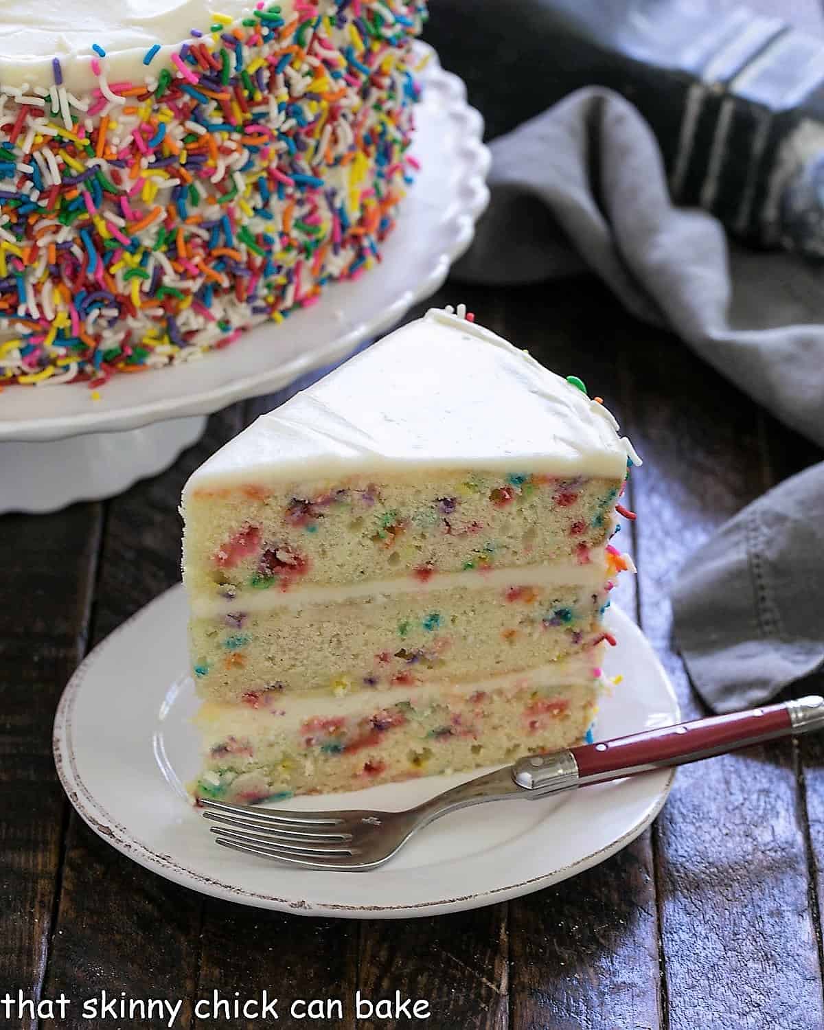 Slice of funfetti cake on a white plate with a red handle fork in front of a cake stand.