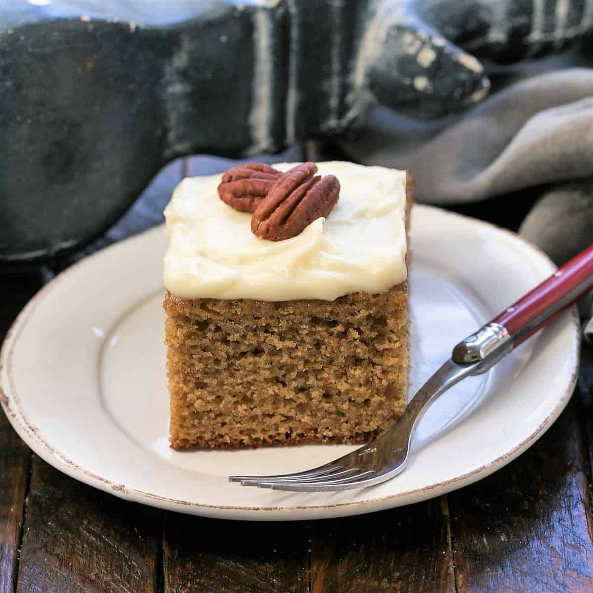 A slice of applesauce spice cake topped with 2 pecan on a round white plate with a red handled fork.