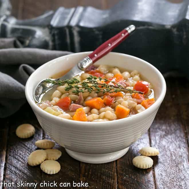 Bowl of bean soup with ham garnished with thyme in a white bowl with a red handle spoon surrounded by oyster crackers